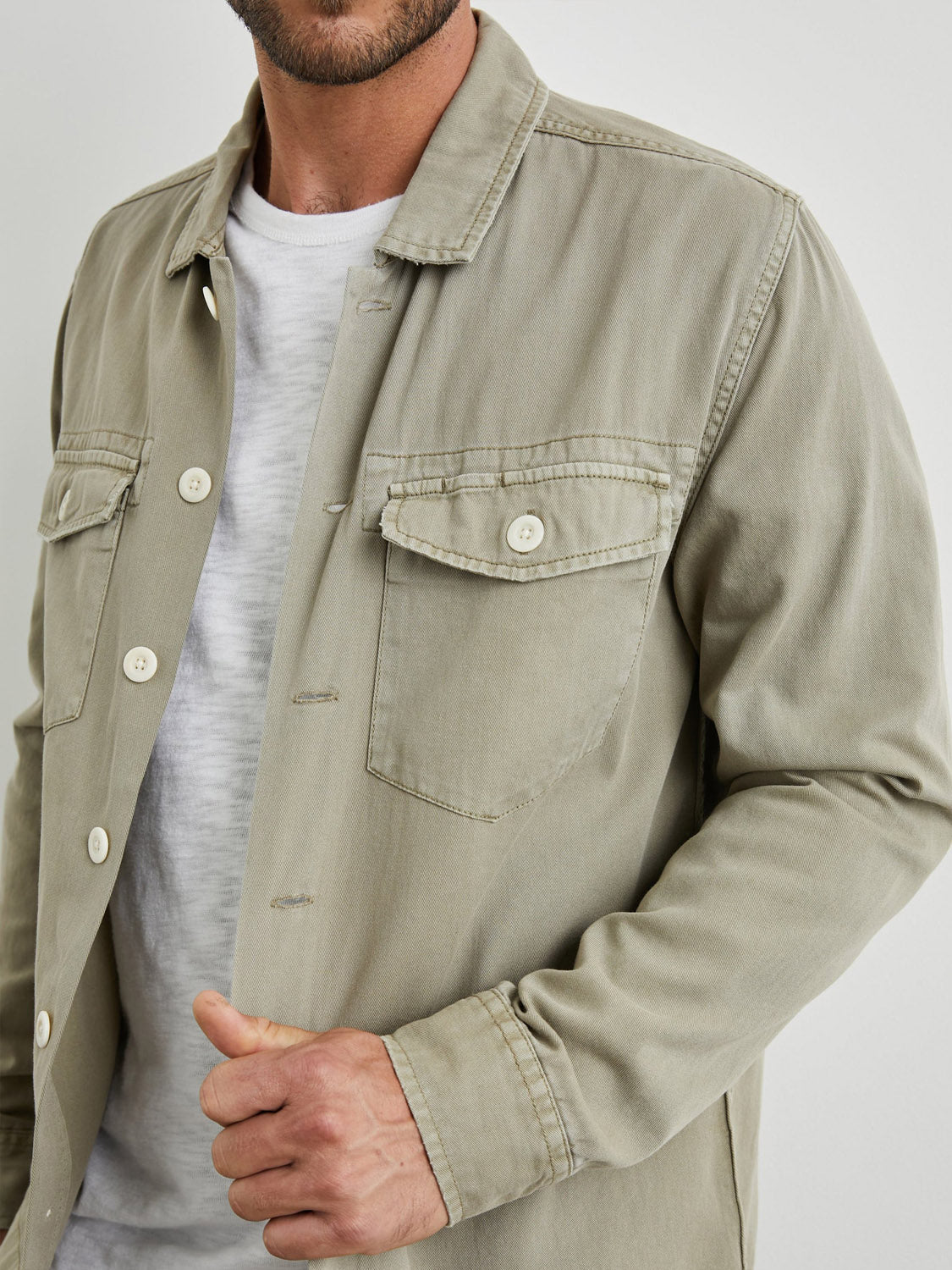 A man wears the Rails Kerouac Shirt Jacket in light beige over a white tee, standing against a plain background. The shirt jacket features two button-flap chest pockets, and he holds its edge with one hand.