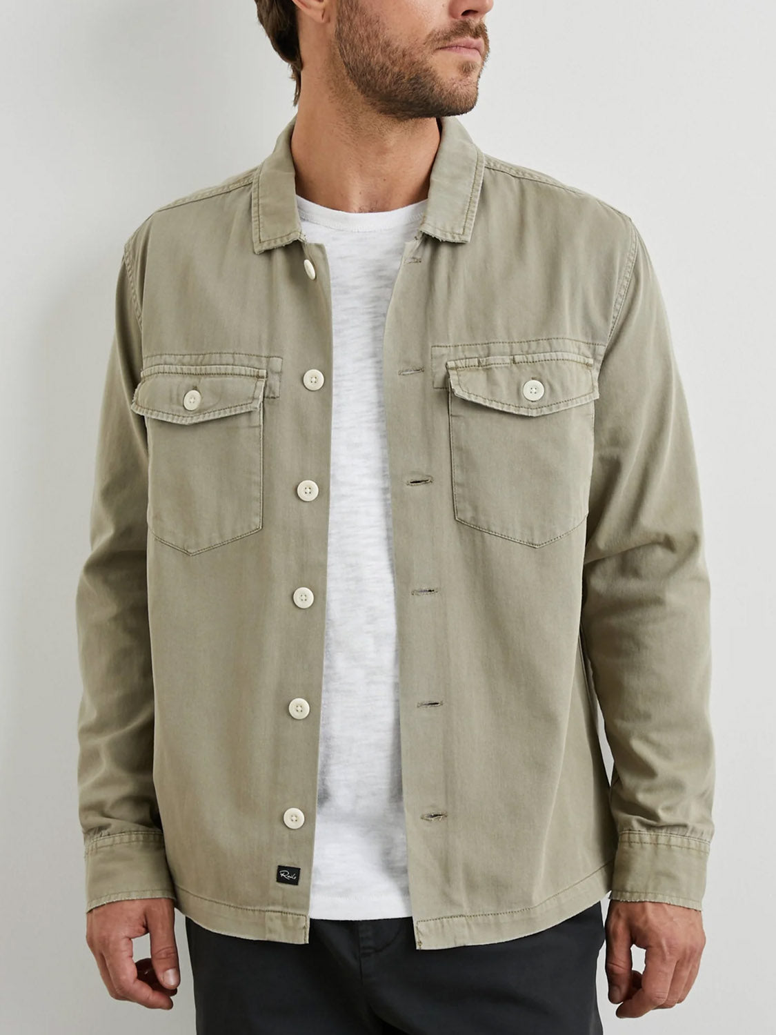 A man wearing the Rails Kerouac Shirt Jacket in light khaki over a white T-shirt stands against a plain white background, his head turned slightly to the right. The shirt jacket features two chest pockets and a military-inspired design.
