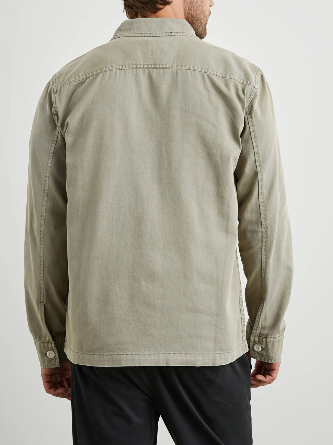 A man is seen from the back wearing the Rails Kerouac Shirt Jacket in light khaki with long sleeves, paired with black pants, set against a plain light background.