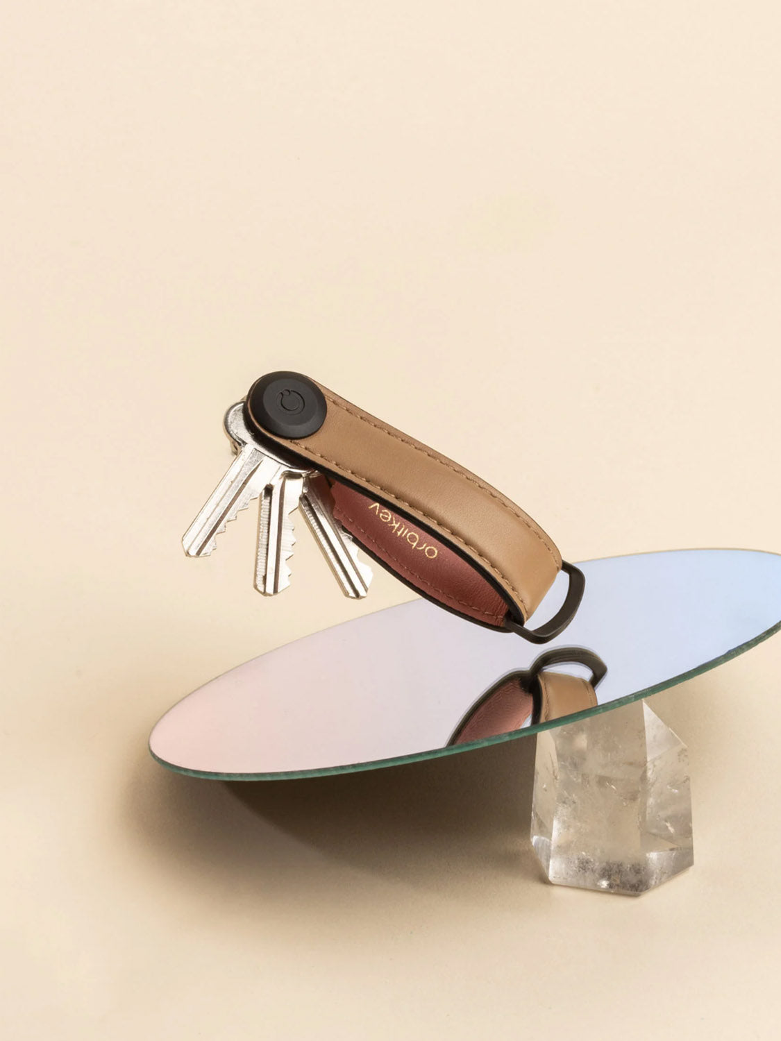 The Orbitkeys Key Organizer Leather, holding multiple silver keys, is displayed on a round mirror set atop a clear, faceted crystal against a beige background.