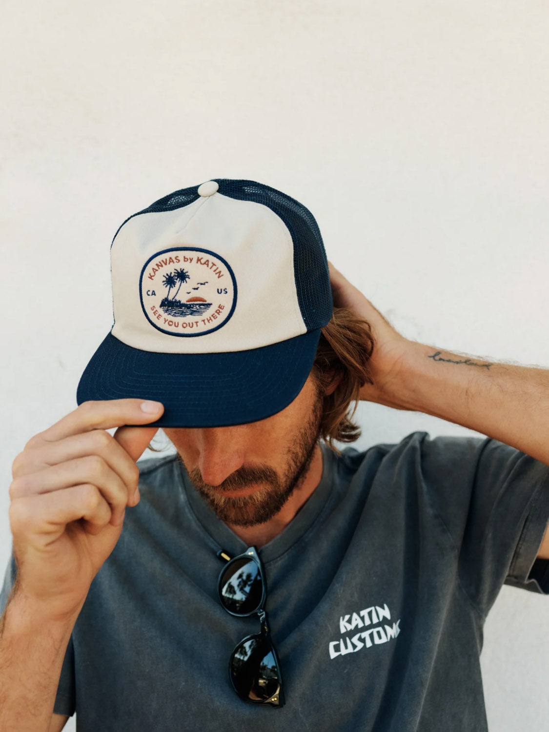 A man in a gray Katin Customs t-shirt adjusts his Katin Keys Trucker Hat with a coastal-themed patch. Featuring cotton twill and a snapback closure, the cap completes his look. Sunglasses hang from his shirt as he stands against a plain light-colored background.