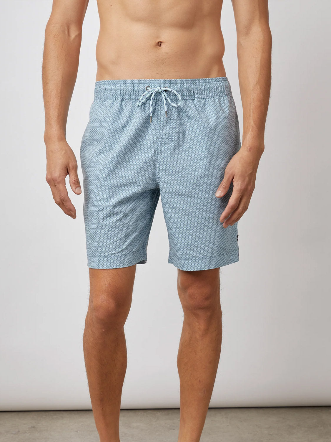 A person stands against a plain background, modeling light blue Rails Kian Swim Shorts with a subtle pattern. Made from a lightweight cotton-blend, these relaxed trunks feature a drawstring fit, while the upper body is partially out of view, emphasizing the casual stance.
