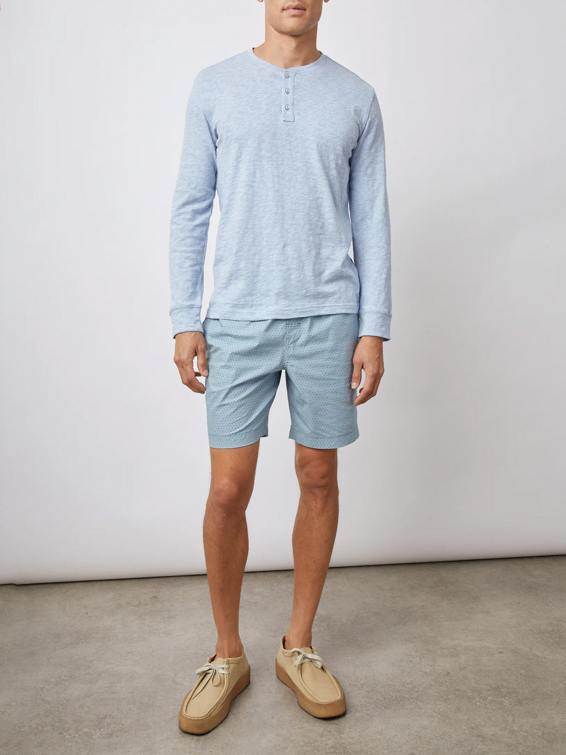 A man wears a light blue long-sleeve henley shirt, Rails Kian Swim Short in a blue pattern, and beige shoes, standing on a concrete floor against a plain white backdrop.