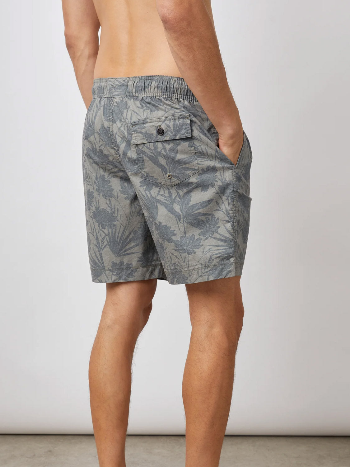 Rear view of a person in gray floral-patterned Rails Kian Swim Shorts, featuring an elastic waistband. The lightweight cotton-blend shorts have a back pocket with a button. They stand shirtless against a plain background, hands in side pockets.