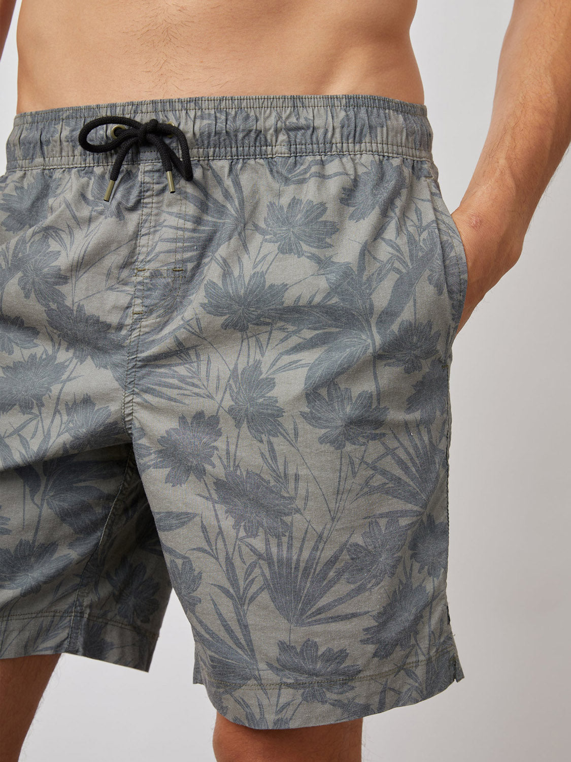 A person in grey floral-patterned Rails Kian Swim Shorts with a black drawstring, hands in pockets. These lightweight cotton-blend shorts by Rails feature a tropical flower and leaf design on an off-white background.
