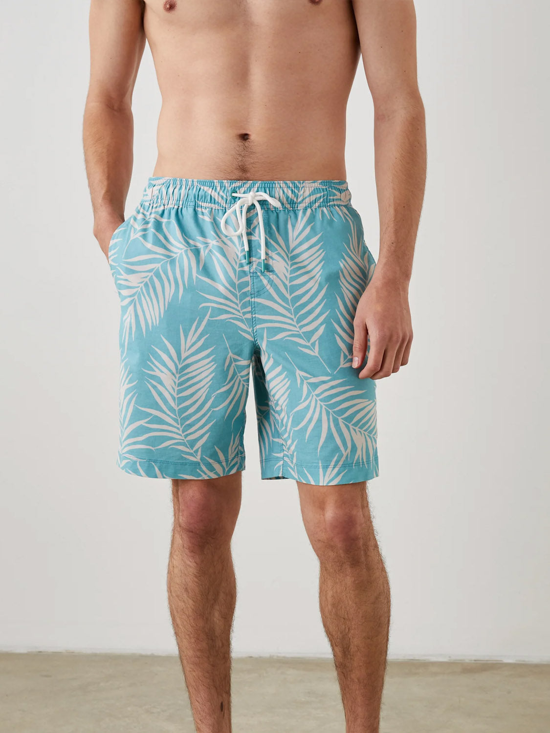 Wearing the Rails Kian Swim Short, a lightweight cotton-blend trunk in blue with white palm leaf patterns, they stand casually with hands in pockets on a light-colored floor against a plain wall. The white drawstring at the waist is prominently displayed.