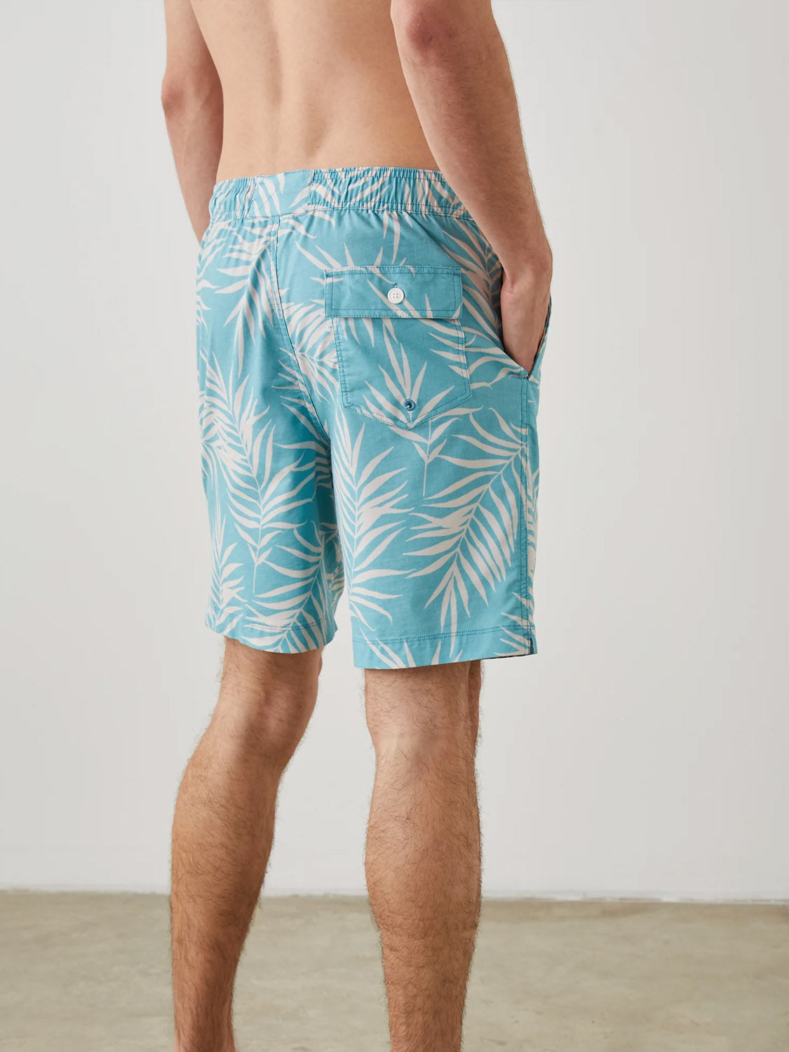 A person stands with their back to the camera, wearing light blue Rails Kian Swim Shorts featuring a white palm leaf design. Made from a lightweight cotton-blend, their right hand rests casually in the pocket while leaning against a plain light-colored wall.