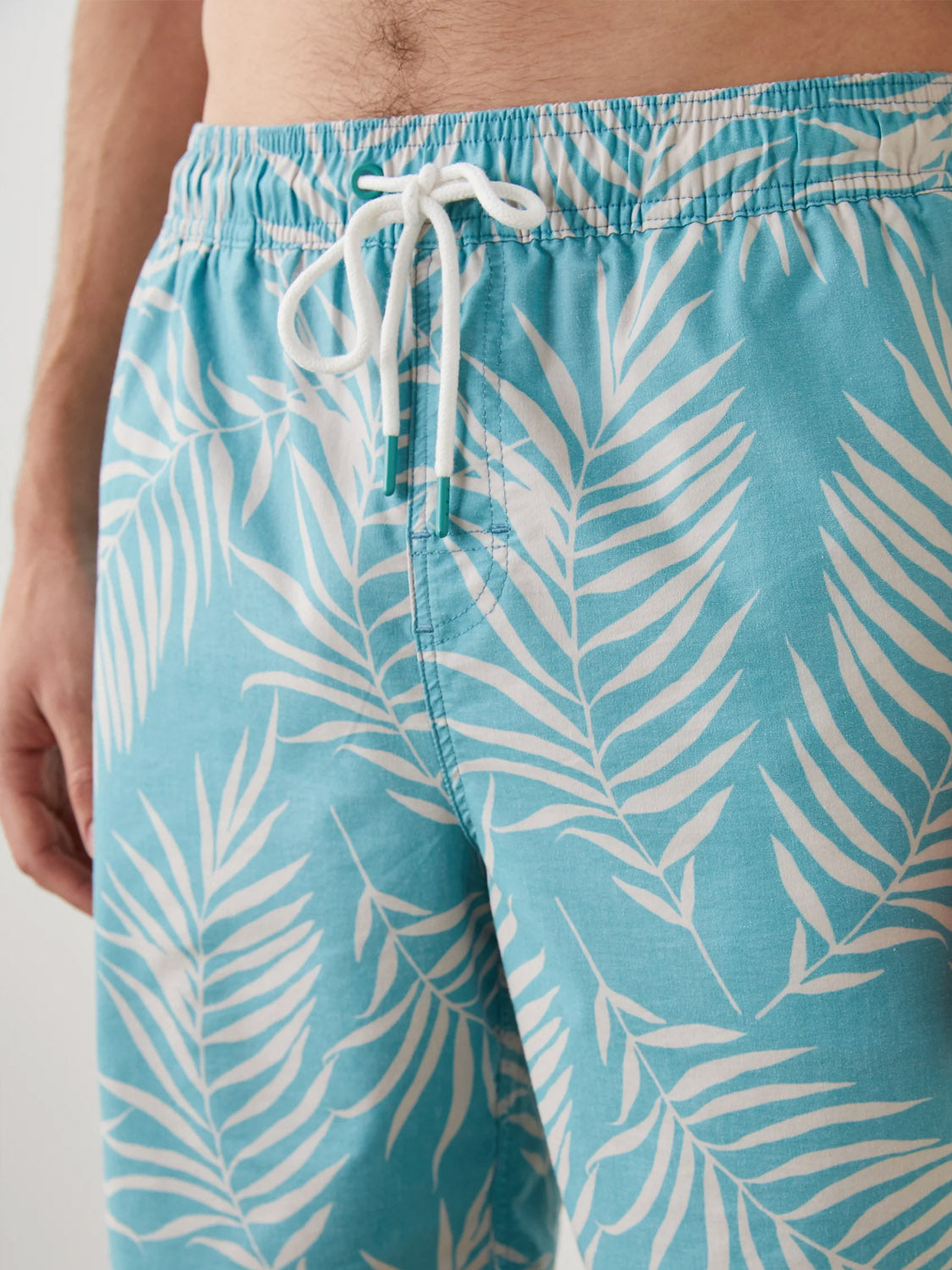 Close-up of a person wearing the Rails Kian Swim Short in aqua blue with a white fern leaf pattern. These lightweight cotton-blend trunks have a white drawstring at the waist, with one hand resting at their side, highlighting the shorts stylish design.