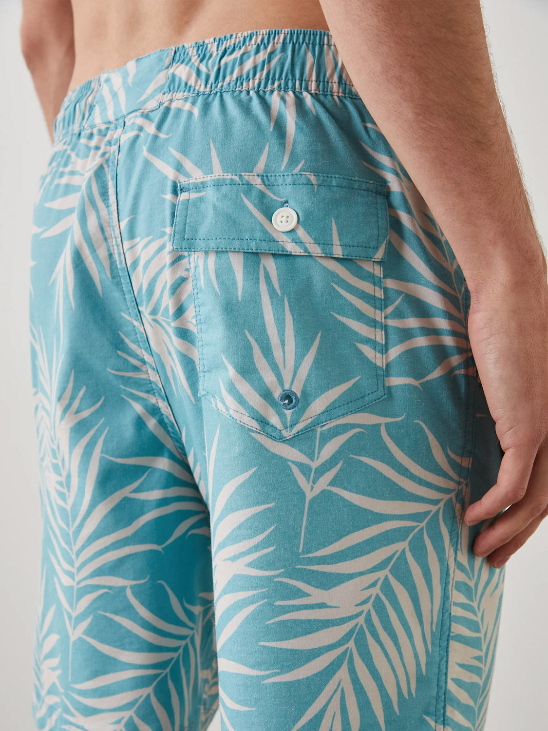 A person wears blue Rails Kian Swim Shorts, featuring white tropical leaf patterns, a buttoned back pocket, and an elastic waistband. Their right hand rests near the pocket of these lightweight cotton-blend shorts.