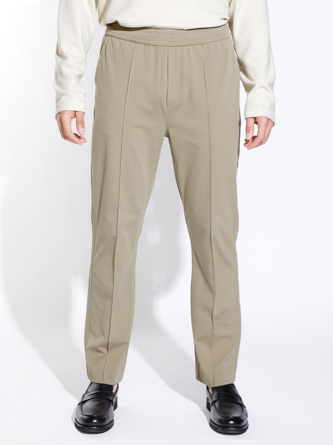 A person wears Civil Society's Klien Loose Fit Dressy Trouser in light beige with an elastic waistband, paired with a cream long-sleeve top and black dress shoes, standing against a plain white background.