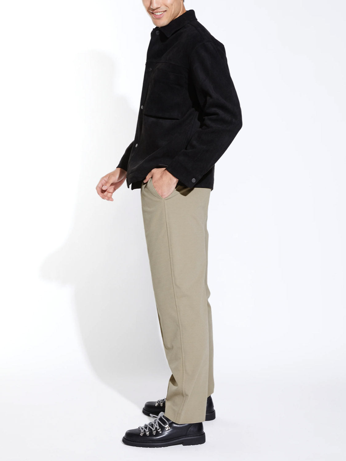 A person stands sideways, smiling with their left hand in their pocket, wearing Civil Society’s Klien Loose Fit Dressy Trouser in beige, a black piqué knit jacket, and black shoes with white laces against a plain white background.