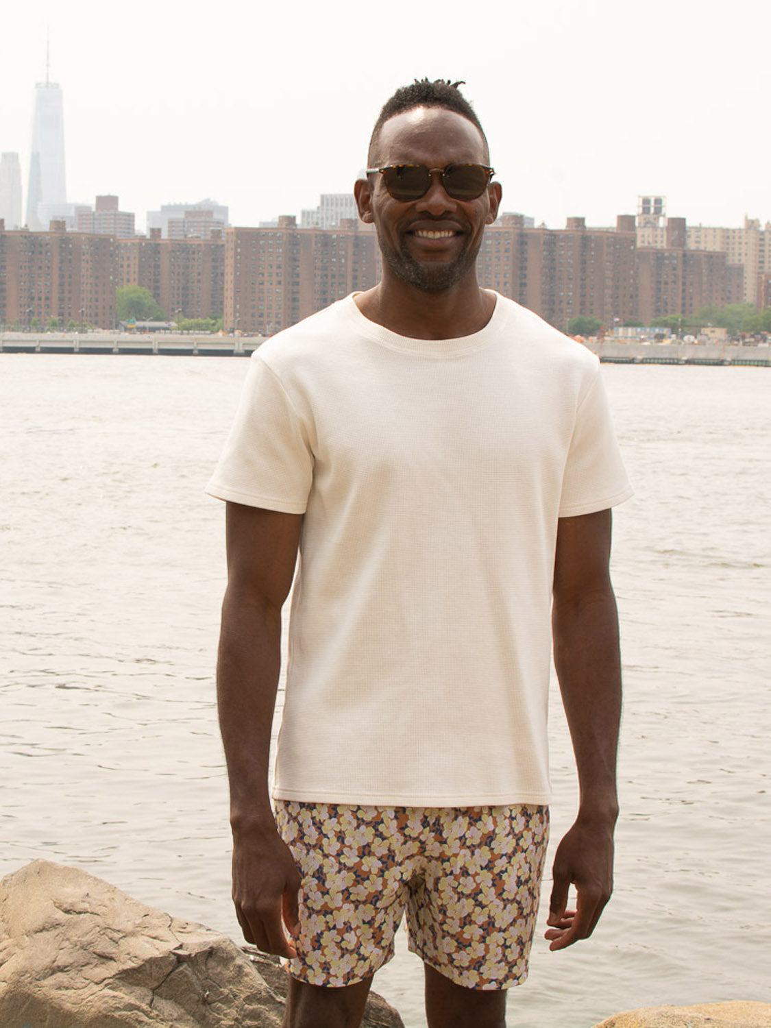 A man in ID Menswear’s Kokio Fast Dry 4-Way Stretch Swim Trunk and sunglasses stands by the water with a city skyline behind him.
