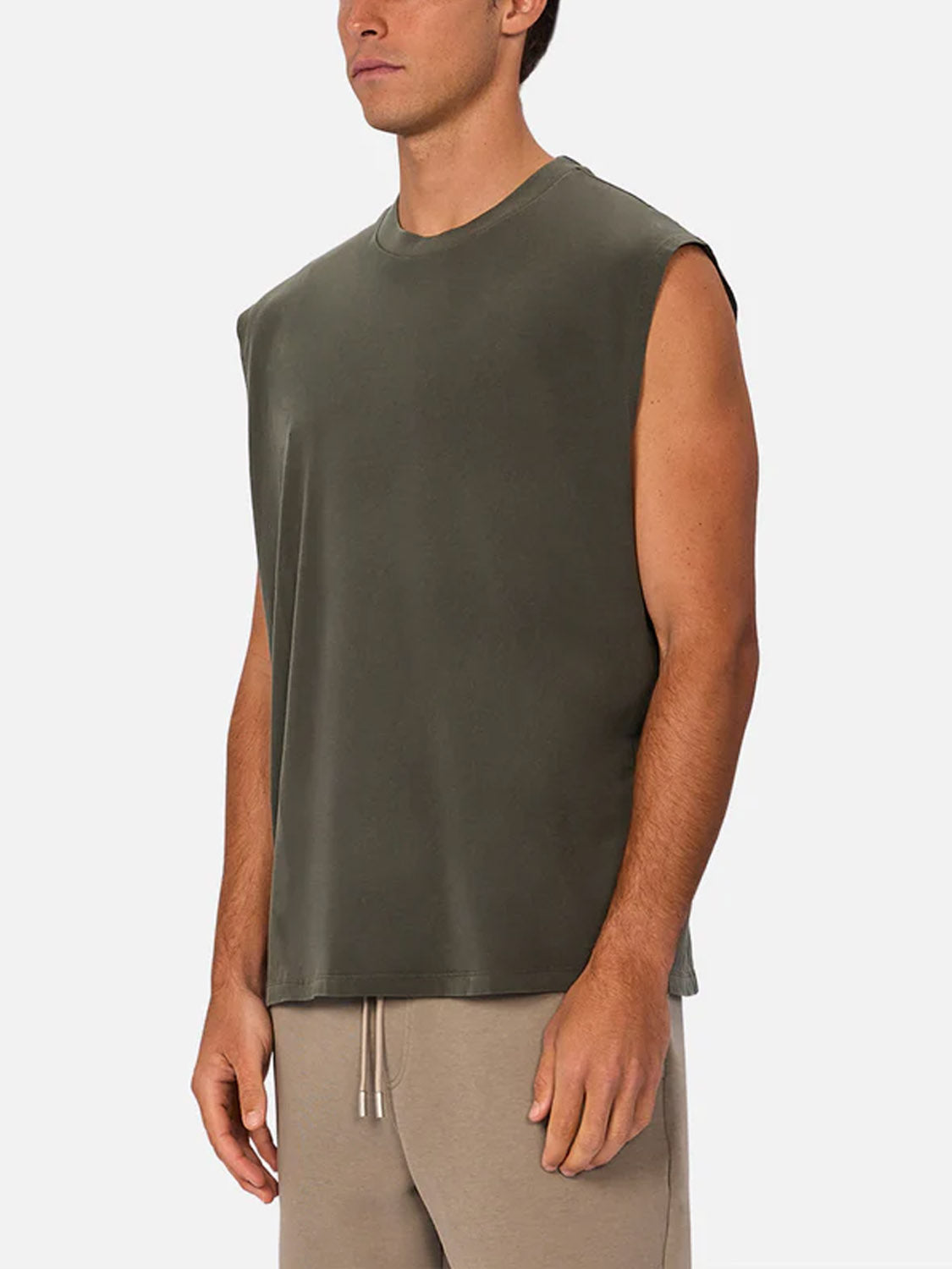 A man stands against a plain white background, turned slightly to the side, wearing Industrie’s The Kosta Sleeveless Tee and beige drawstring pants.