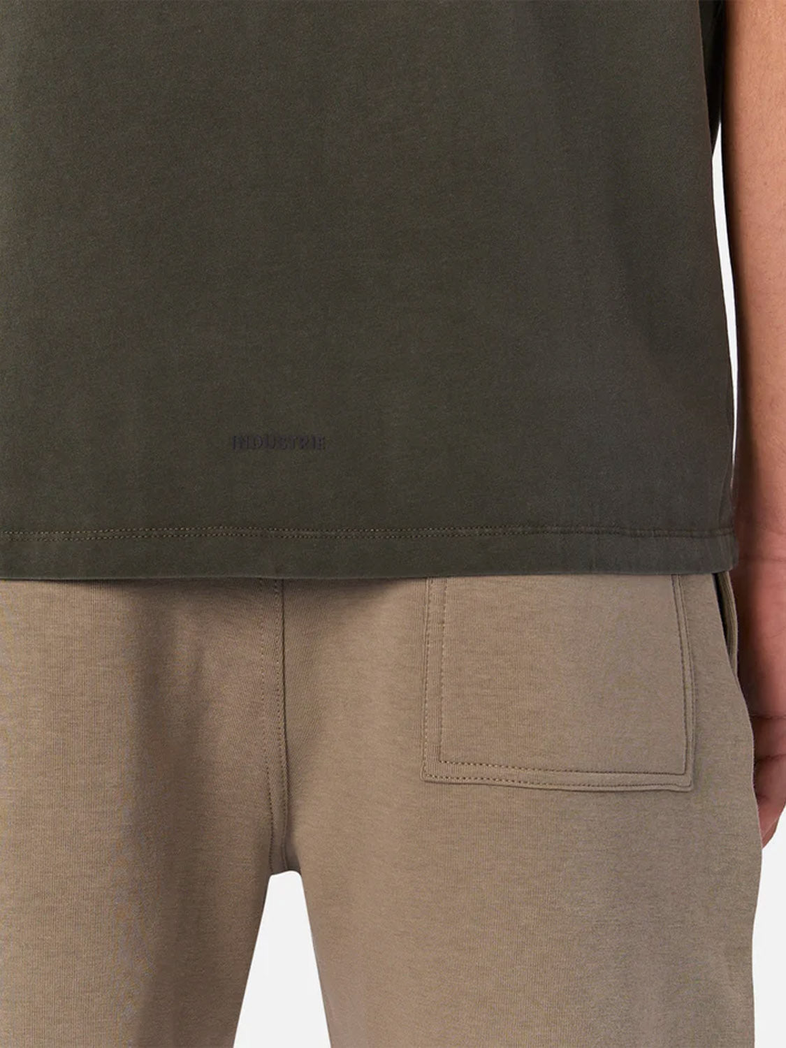 A person shown from waist to mid-thigh wears Industrie’s The Kosta Sleeveless Tee in dark green, paired with light brown pants featuring a square patch pocket on the thigh.