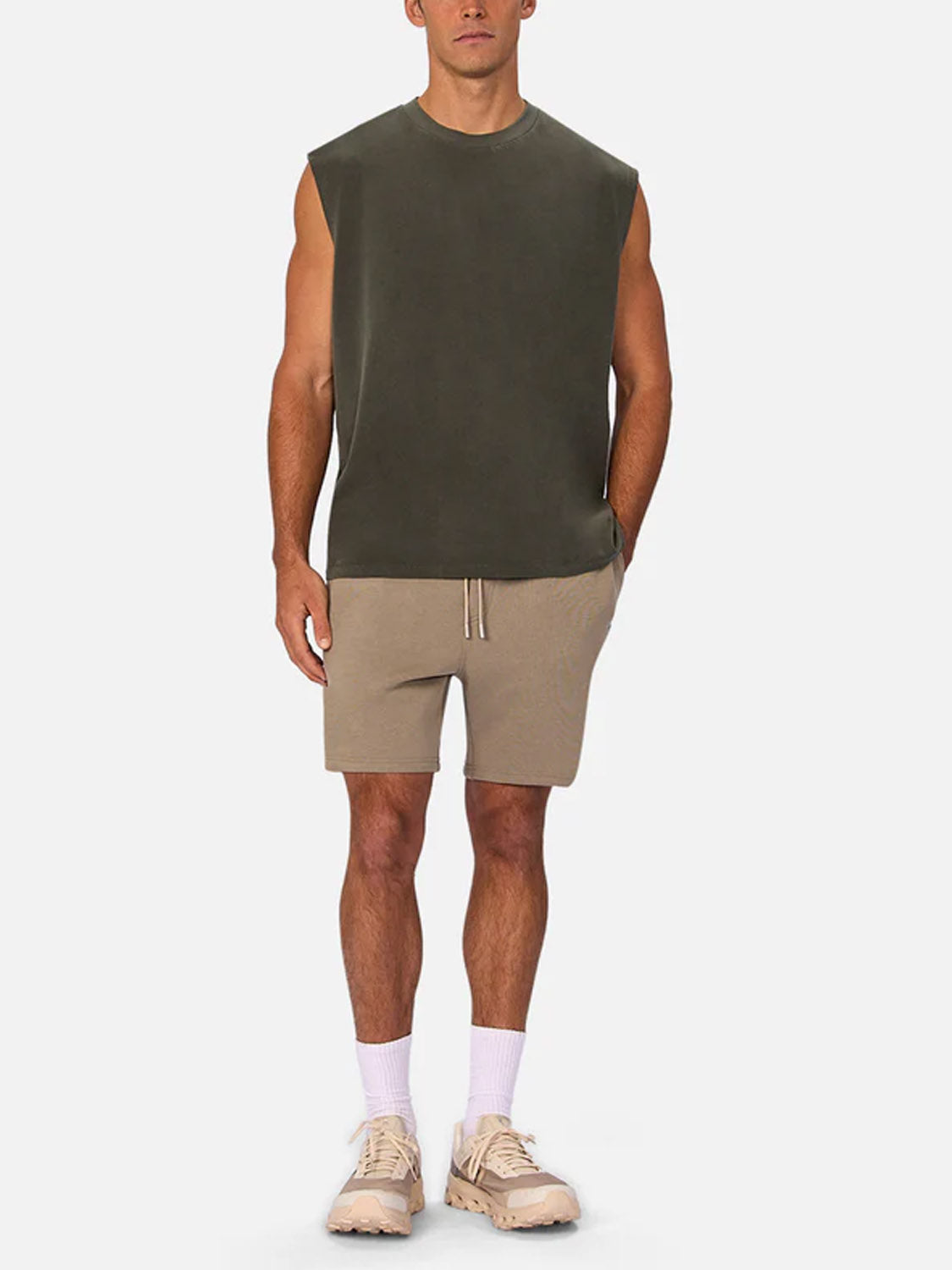 A man faces forward against a white background wearing Industrie’s The Kosta Sleeveless Tee in dark green, paired with beige shorts, white socks, and beige sneakers.