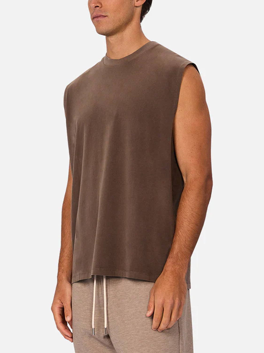 A man models The Kosta Sleeveless Tee by Industrie, paired with light beige drawstring pants. He stands in front of a plain light background, turned slightly to the side with his arms relaxed at his sides.