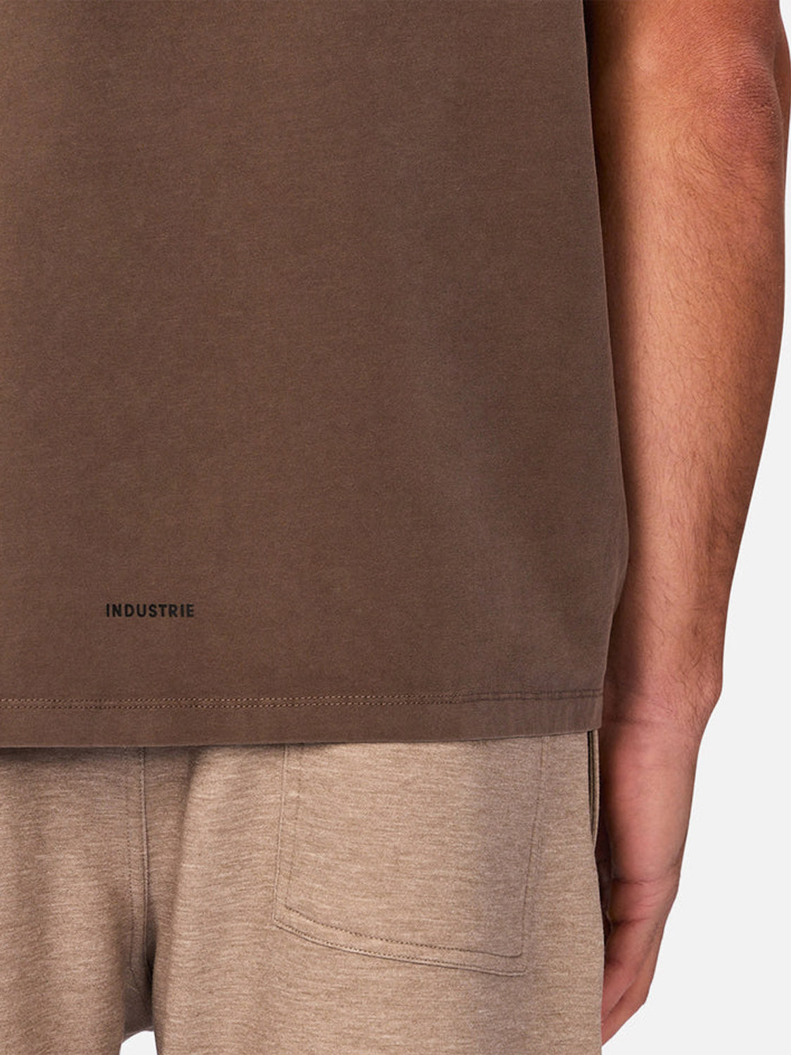 A close-up of a person in the Kosta Sleeveless Tee by Industrie, a brown quick-dry athletic shirt with "INDUSTRIE" printed near the bottom hem. Beige pants are also visible, as is the wearer’s right arm.