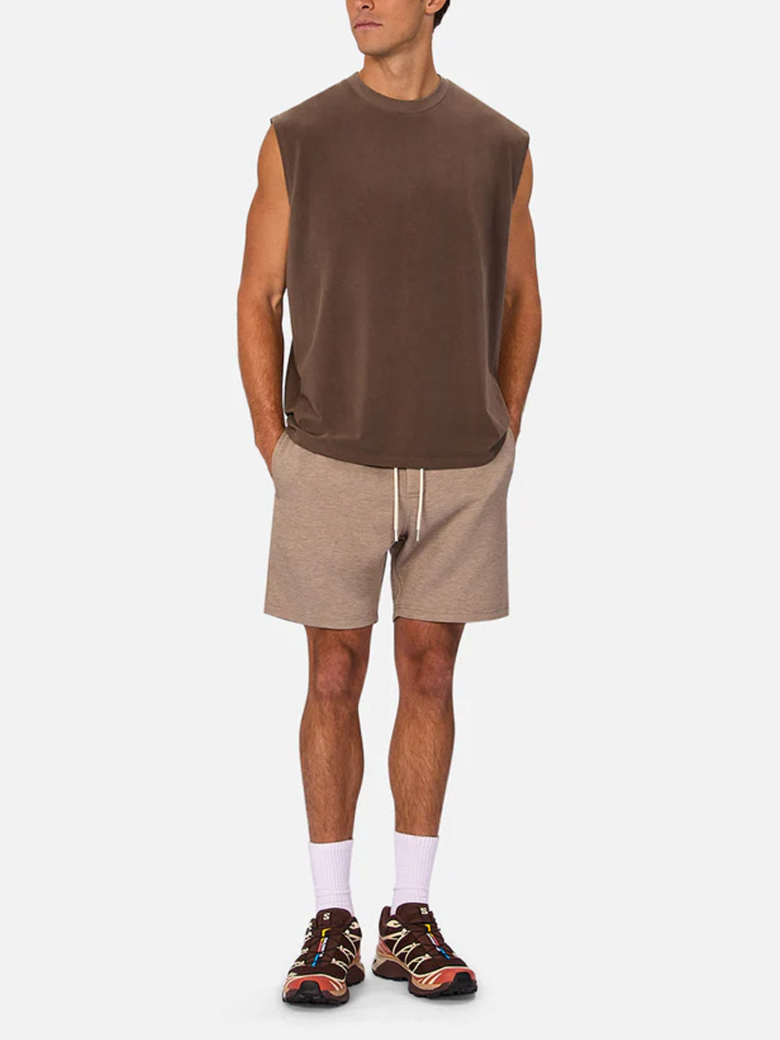 A man poses with hands in his pockets, wearing Industrie’s The Kosta Sleeveless Tee, beige drawstring shorts, white socks, and multicolored sneakers against a plain white background.