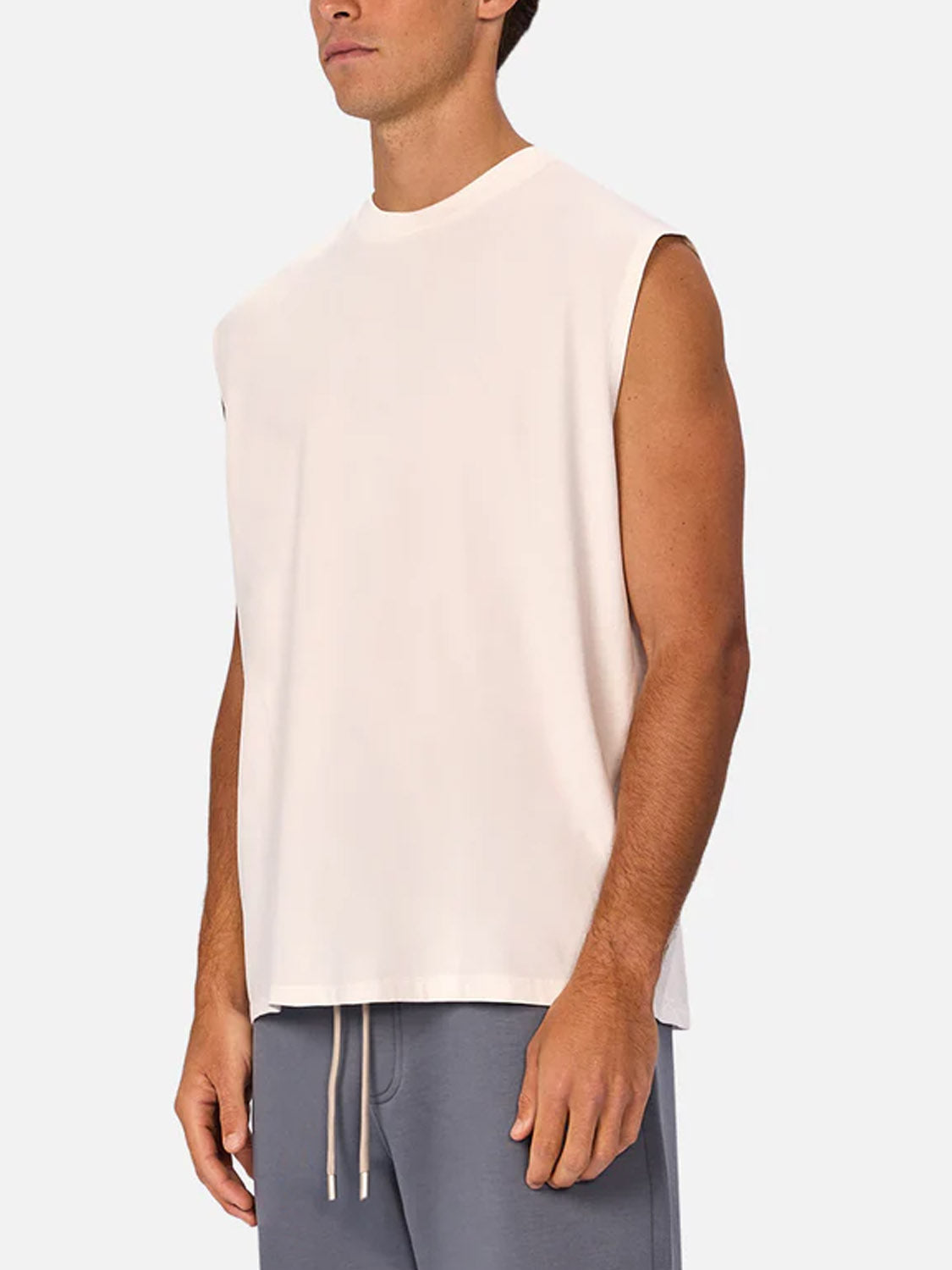 A man wears the Industrie Kosta Sleeveless Tee in light beige with gray drawstring shorts, standing against a plain light background. His face is partially out of frame.