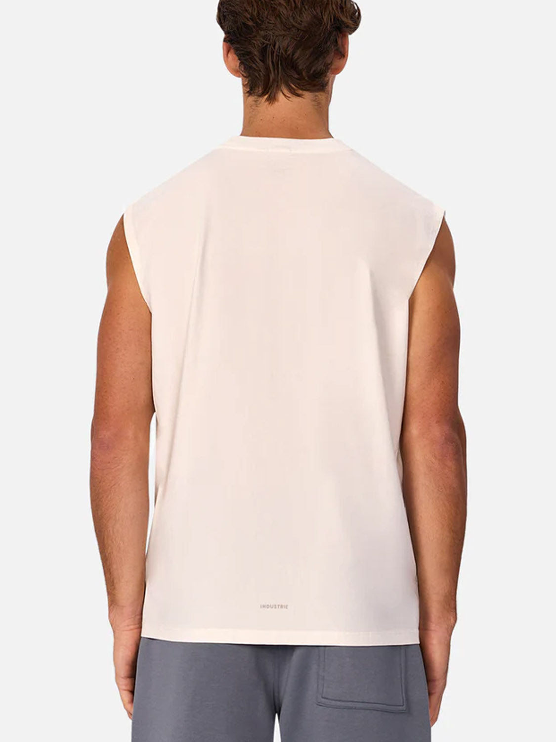 A man is seen from the back wearing The Kosta Sleeveless Tee by Industrie in off-white, paired with gray shorts featuring a visible back pocket, standing against a plain light background.