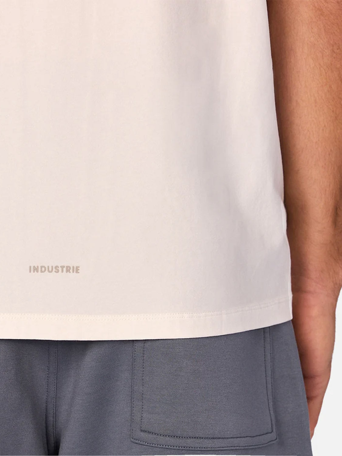 A close-up of a person wearing The Kosta Sleeveless Tee by Industrie in light beige and gray pants, showing only the lower half of the shirt with "Industrie" near the hem and the upper part of the pants from the back.