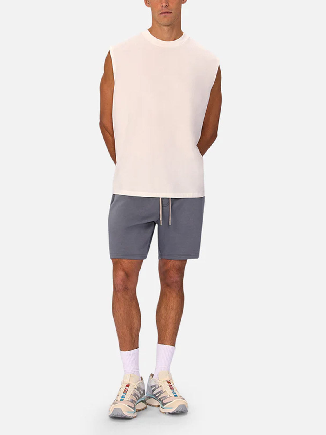 A man stands facing forward in the Industrie Kosta Sleeveless Tee, paired with gray shorts, white socks, and beige sneakers. His arms are behind his back against a plain light gray background.