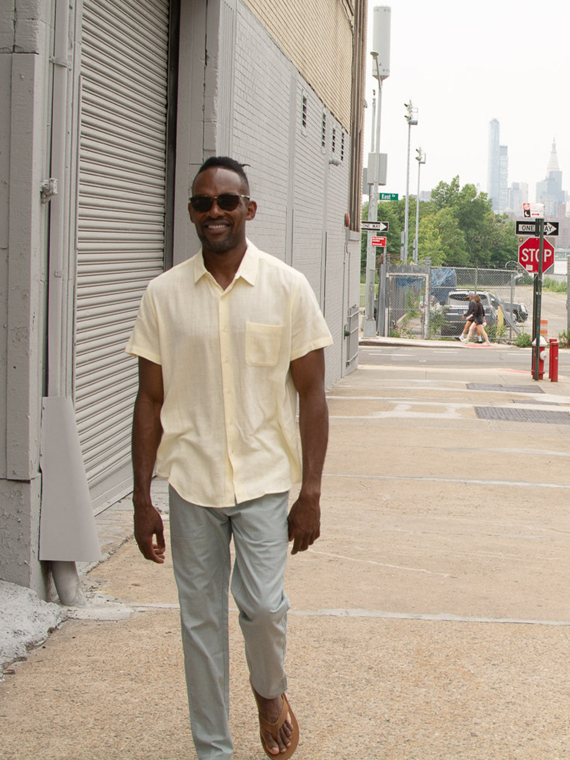 A man in sunglasses, a light yellow short-sleeve shirt, ID Menswear’s Kruger Relaxed Pant, and sandals strolls a city sidewalk by a gray building, enjoying warm-weather comfort amid urban scenery and blurred passersby.