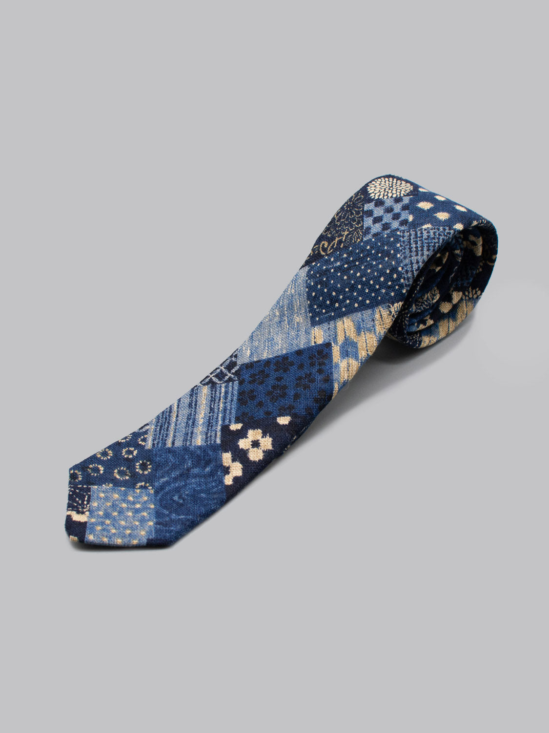 The Japanese Printed Kyoto Tie by ID Menswear, handmade in Brooklyn, features blue patchwork with florals, stripes, and geometric designs inspired by Japanese prints. Shown on a light gray background—ideal for business formal wear.