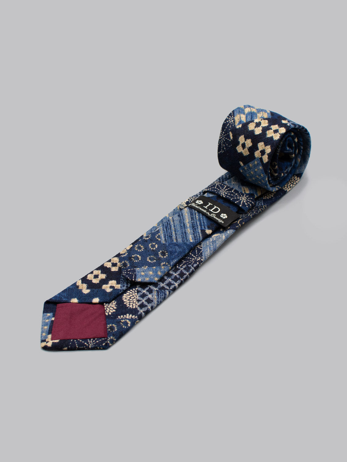 The Japanese Printed Kyoto Tie by ID Menswear showcases blue Japanese patterns with cream geometric and floral designs, a maroon tip, and black label. Handmade in Brooklyn, its ideal for business formal occasions. Shown partially rolled on a light gray background.
