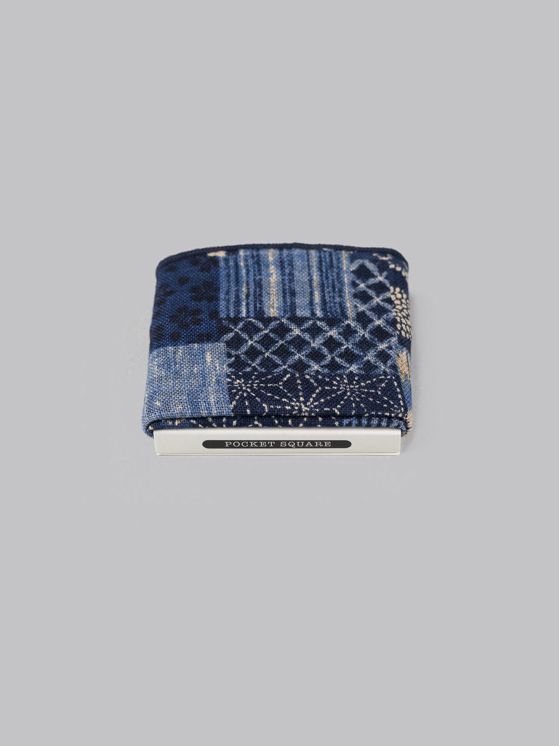 The Kyoto Pocket Square by ID Menswear is a blue patterned cotton accessory with geometric designs, handmade in Brooklyn, neatly folded and held by a silver POCKET SQUARE clip, shown on a plain gray background.