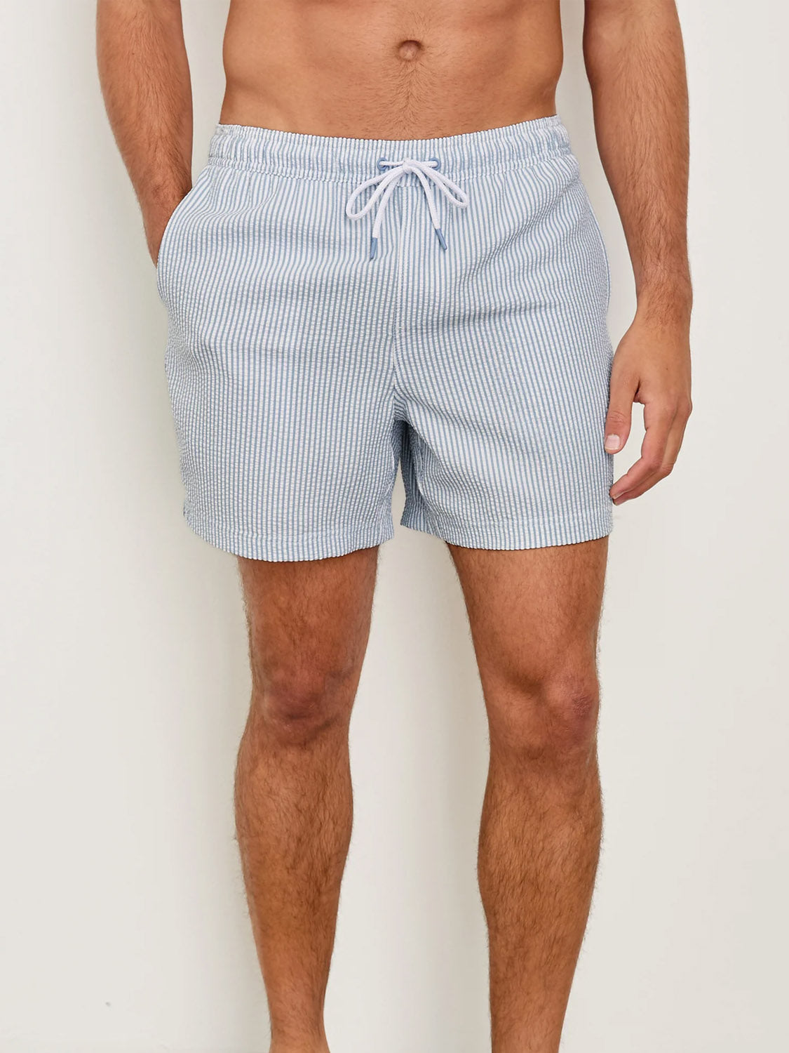 A person stands against a plain background with hands in pockets, showcasing the Rails La Brea Swim Short—a light blue and white vertically striped polyester seersucker style with an adjustable drawstring waist. Only the lower torso and upper legs are shown.