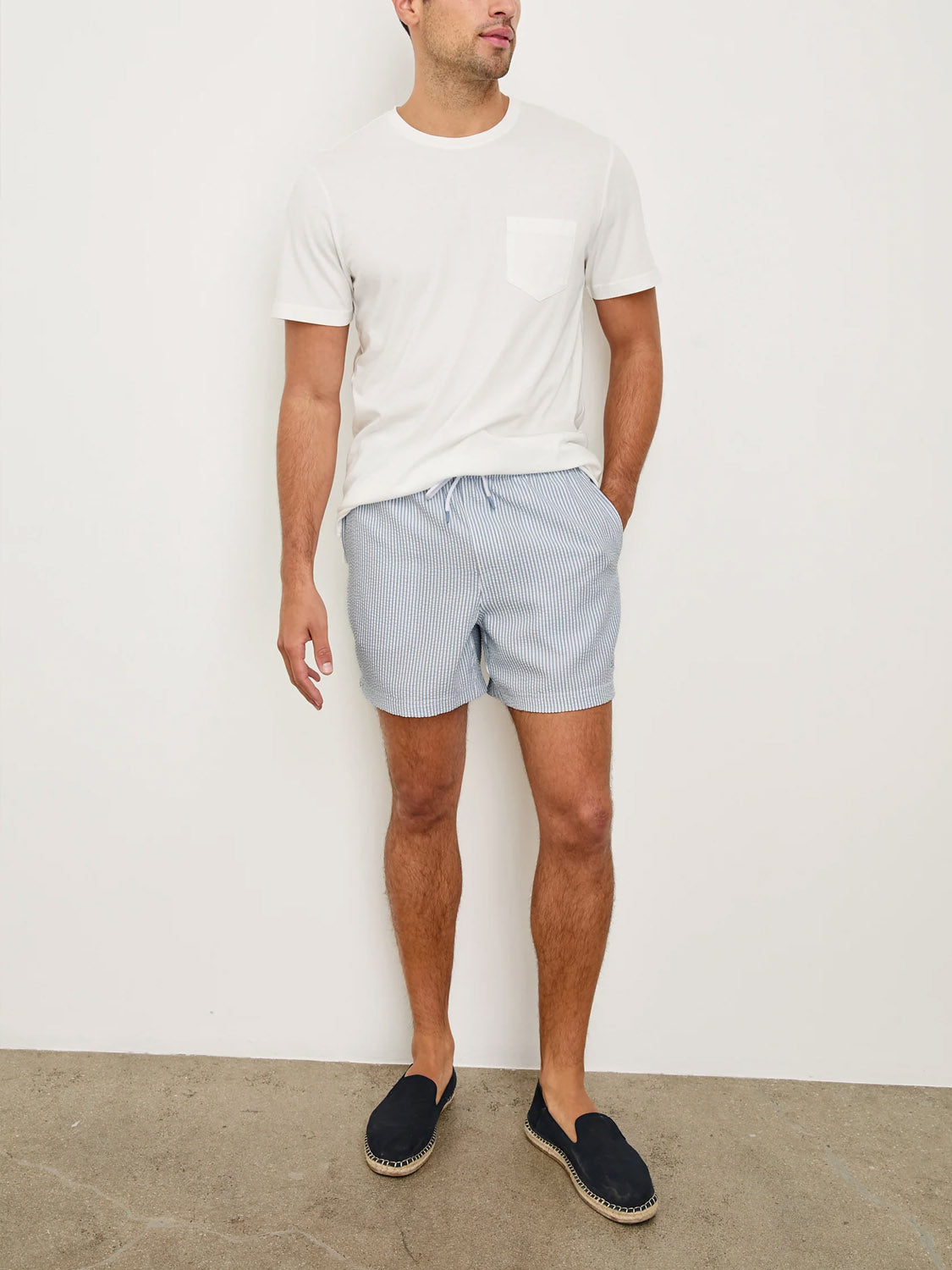 A man stands against a white wall wearing Rails La Brea Swim Short, a light blue striped polyester seersucker style with an adjustable drawstring, paired with a white T-shirt and dark slip-on shoes. He looks slightly left with hands at his sides.