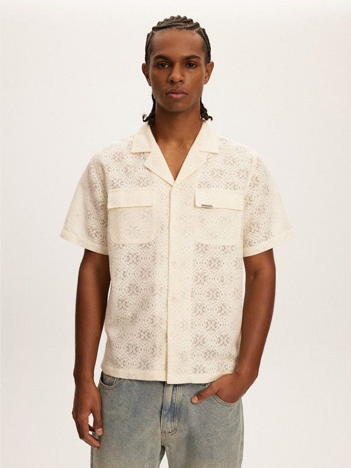 With braided hair and coastal vibes, a person stands against a plain background wearing a short-sleeved Kuwalla Lace Yacht Shirt. This cream-colored, collared shirt has two chest pockets and is perfectly paired with lightweight light blue jeans.