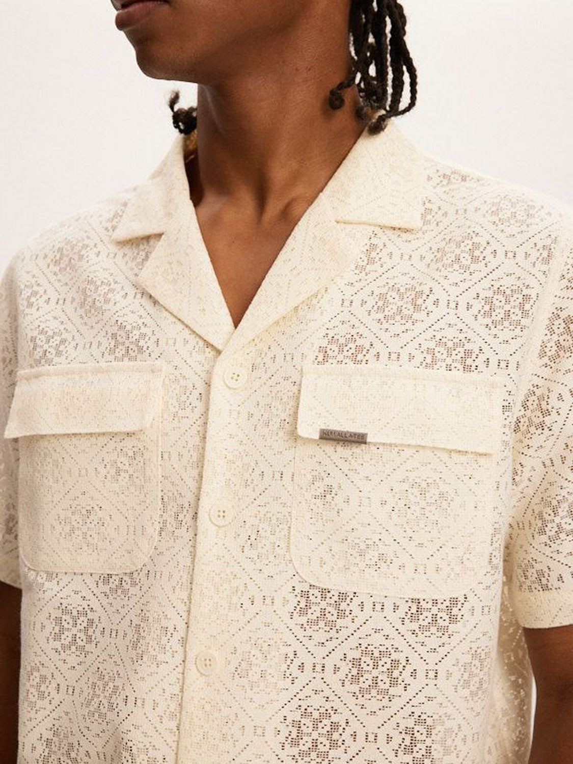 A person with braided hair is shown from the neck down wearing a Kuwalla Lace Yacht Shirt in cream. This lightweight short-sleeve shirt has a collar, pockets, and a diamond design that exudes coastal vibes.