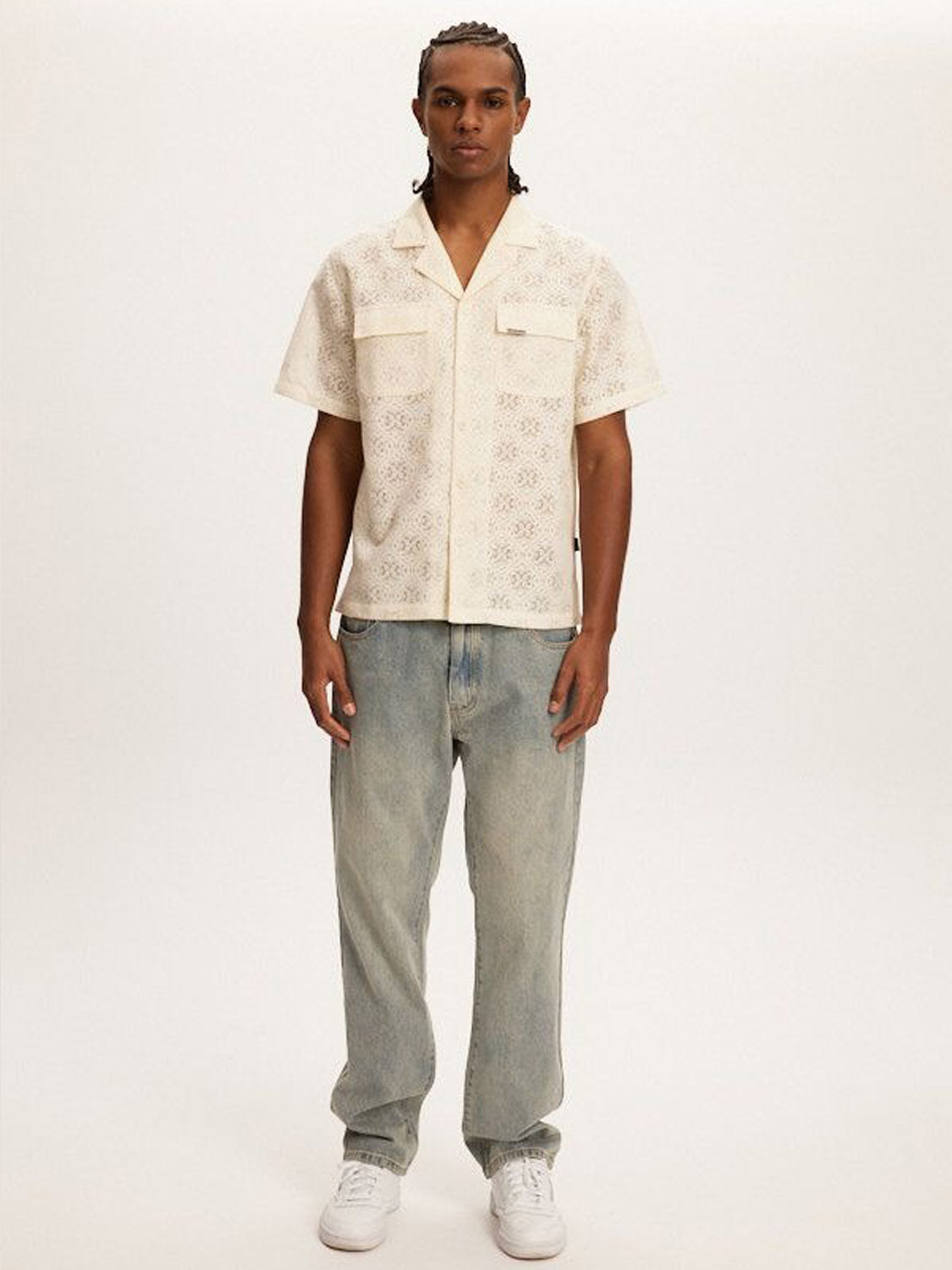 A person with braided hair, embodying coastal vibes, stands against a plain backdrop wearing a light cream patterned Kuwalla Lace Yacht Shirt, light blue baggy jeans, and white sneakers, displaying a relaxed posture and neutral expression.