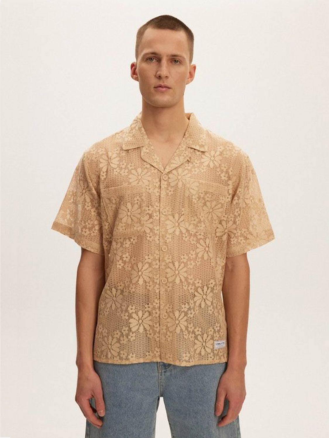 In a minimalist setting, someone showcases the Kuwalla Lace Yacht Shirt, a beige, short-sleeved shirt with a classic fit and floral pattern. Its lightweight fabric complements their short hair and blue jeans seamlessly.