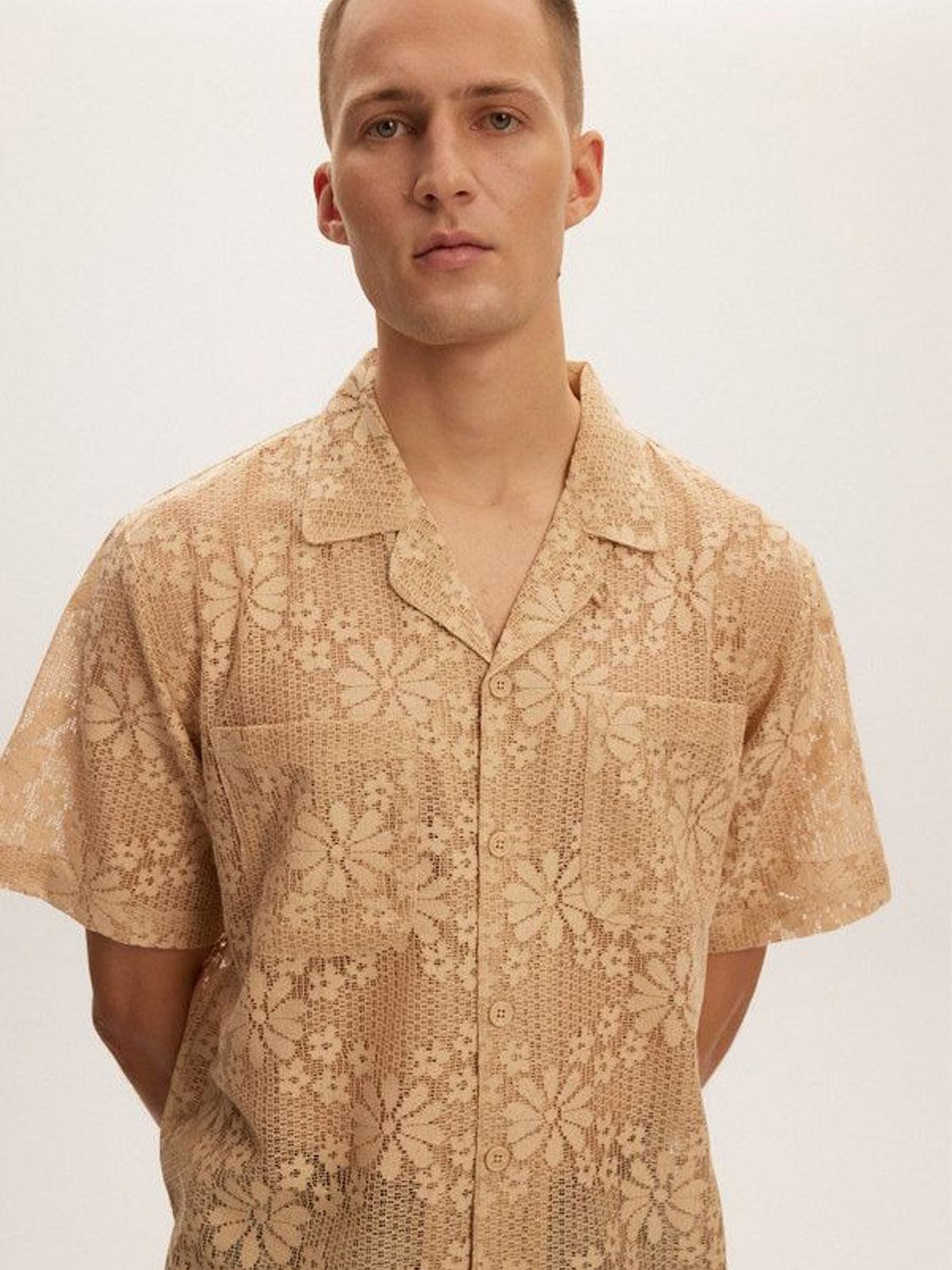 A man wears a Kuwalla Lace Yacht Shirt with a floral pattern, made from lightweight fabric. The brands beige, short-sleeved classic fit design pairs with the plain backdrop as he stands neutrally with his hands behind his back.