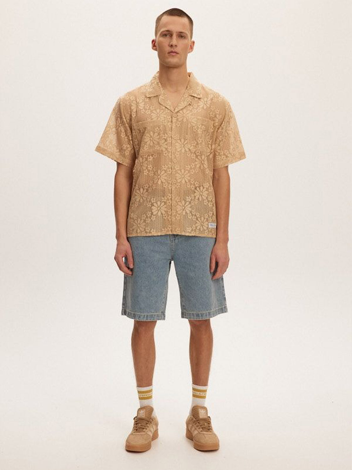 A man stands facing forward against a white background, wearing the Kuwalla Lace Yacht Shirt by Kuwalla, lightweight and beige, paired with knee-length denim shorts, beige socks with yellow stripes, and beige sneakers—ideal for casual days.