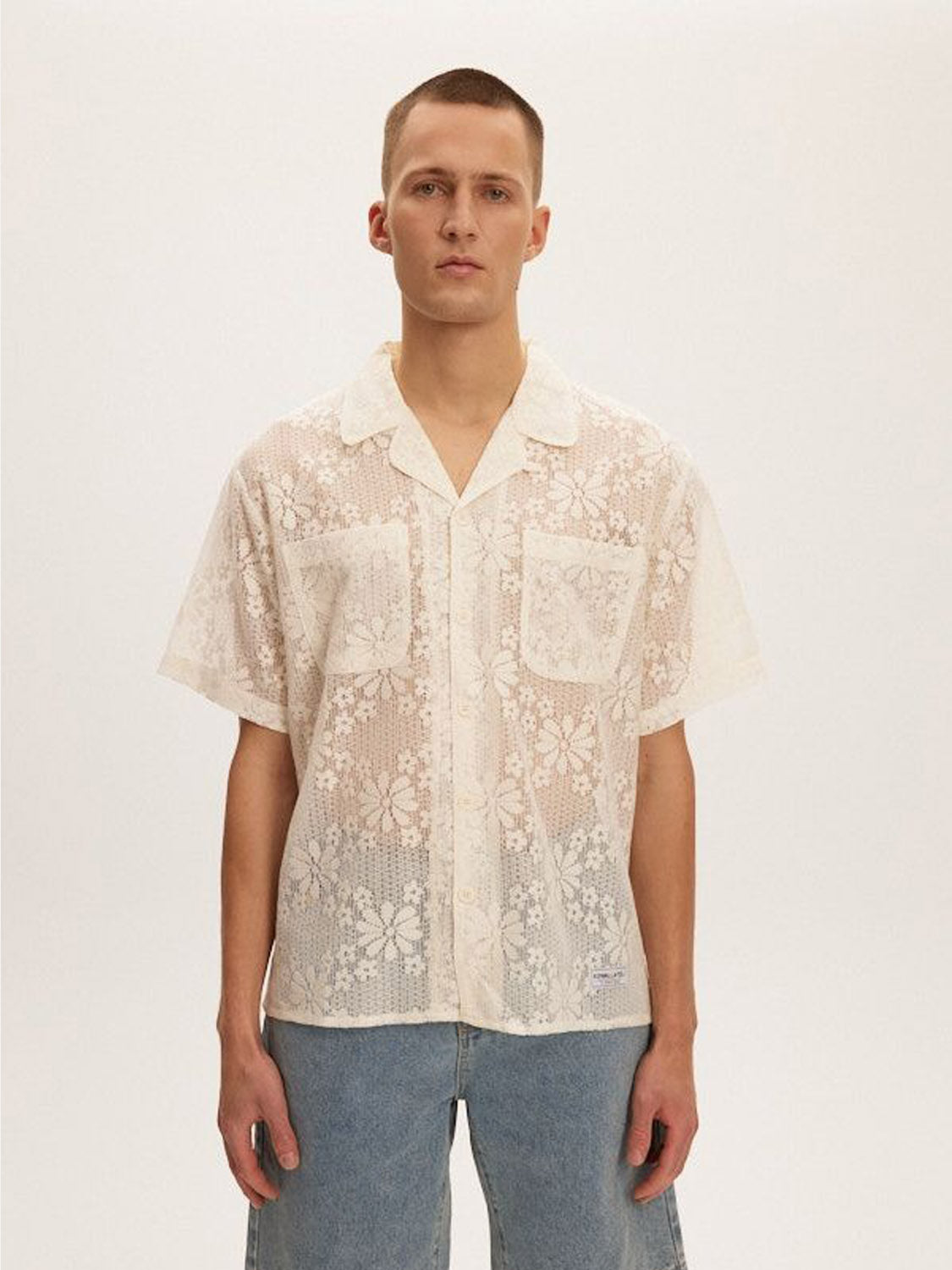 A person faces the camera wearing a sheer Kuwalla Lace Yacht Shirt featuring floral patterns over a plain white undershirt. The classic fit pairs well with their light blue jeans and short hair, standing out against the plain white background thanks to its lightweight fabric.
