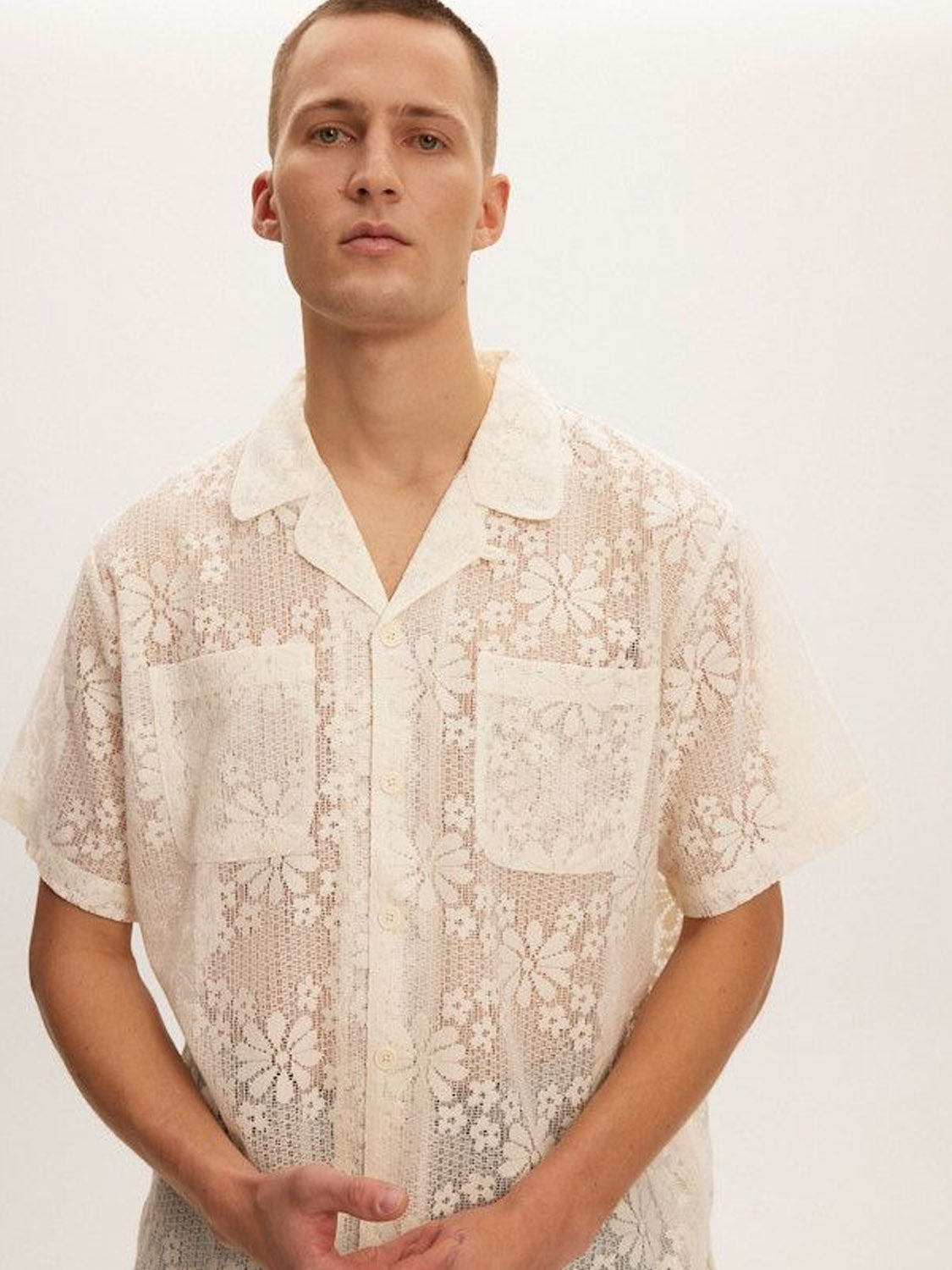 A person wears a Kuwalla Lace Yacht Shirt, a short-sleeve, cream shirt made from lightweight fabric with sheer lace and delicate floral patterns, set against a plain white background.