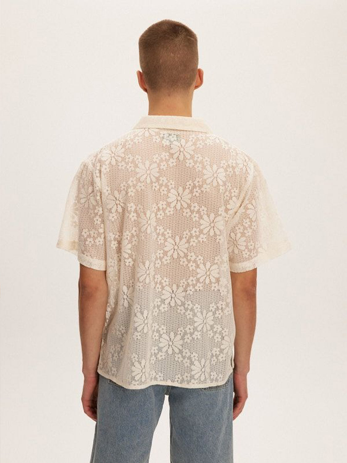 A person with short hair stands away, wearing the Kuwalla Lace Yacht Shirt in beige and light blue jeans made of lightweight fabric against a plain white background.