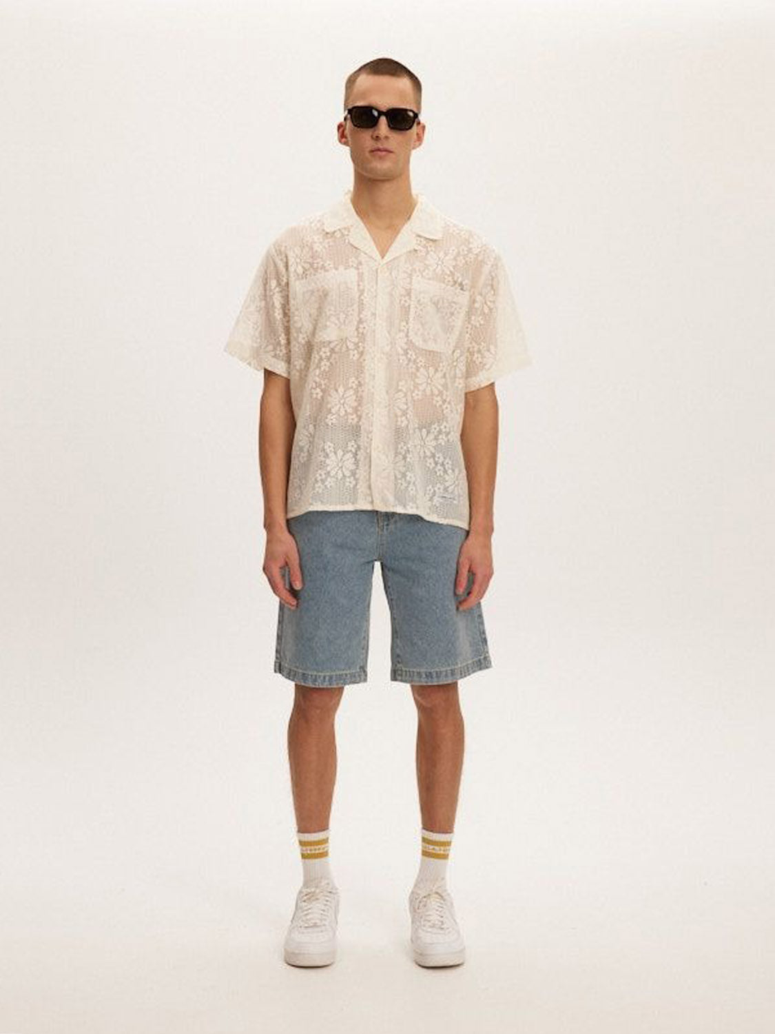 A person in sunglasses, a Kuwalla Lace Yacht Shirt, denim shorts, white socks with yellow stripes, and white sneakers stands against a plain background.