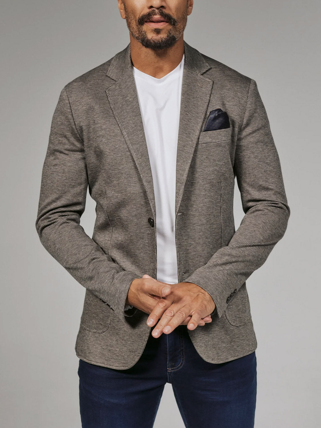 A man models the 7 Diamonds Lafayette Blazer by 7 Diamond over a white tee and blue jeans, standing against a plain background with his hands loosely clasped in front.