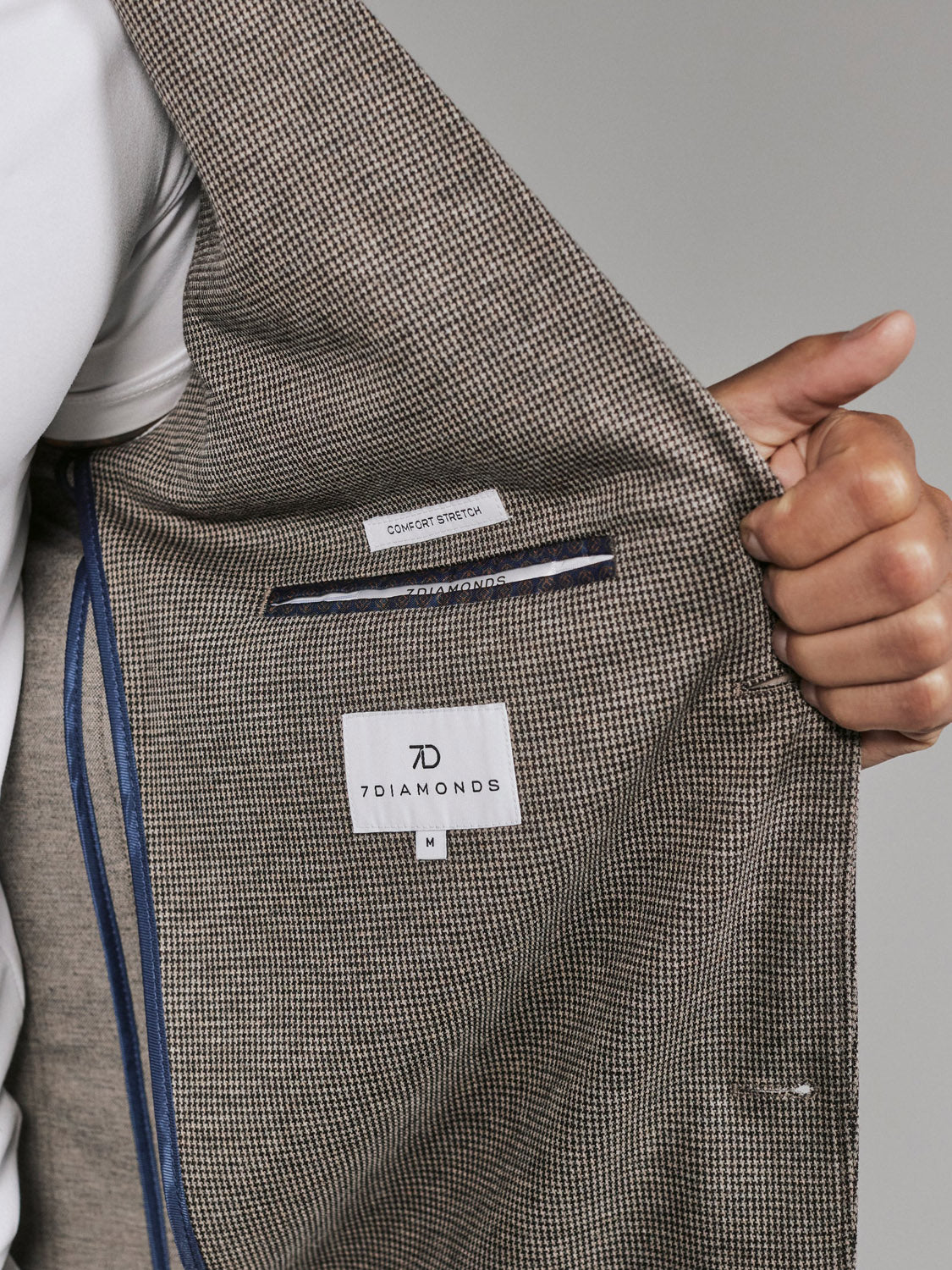 A person opens the 7 Diamonds Lafayette Blazer, revealing its label tags—7 Diamond and Comfort Stretch—and a blue-accented inner pocket, all set against a neutral background.