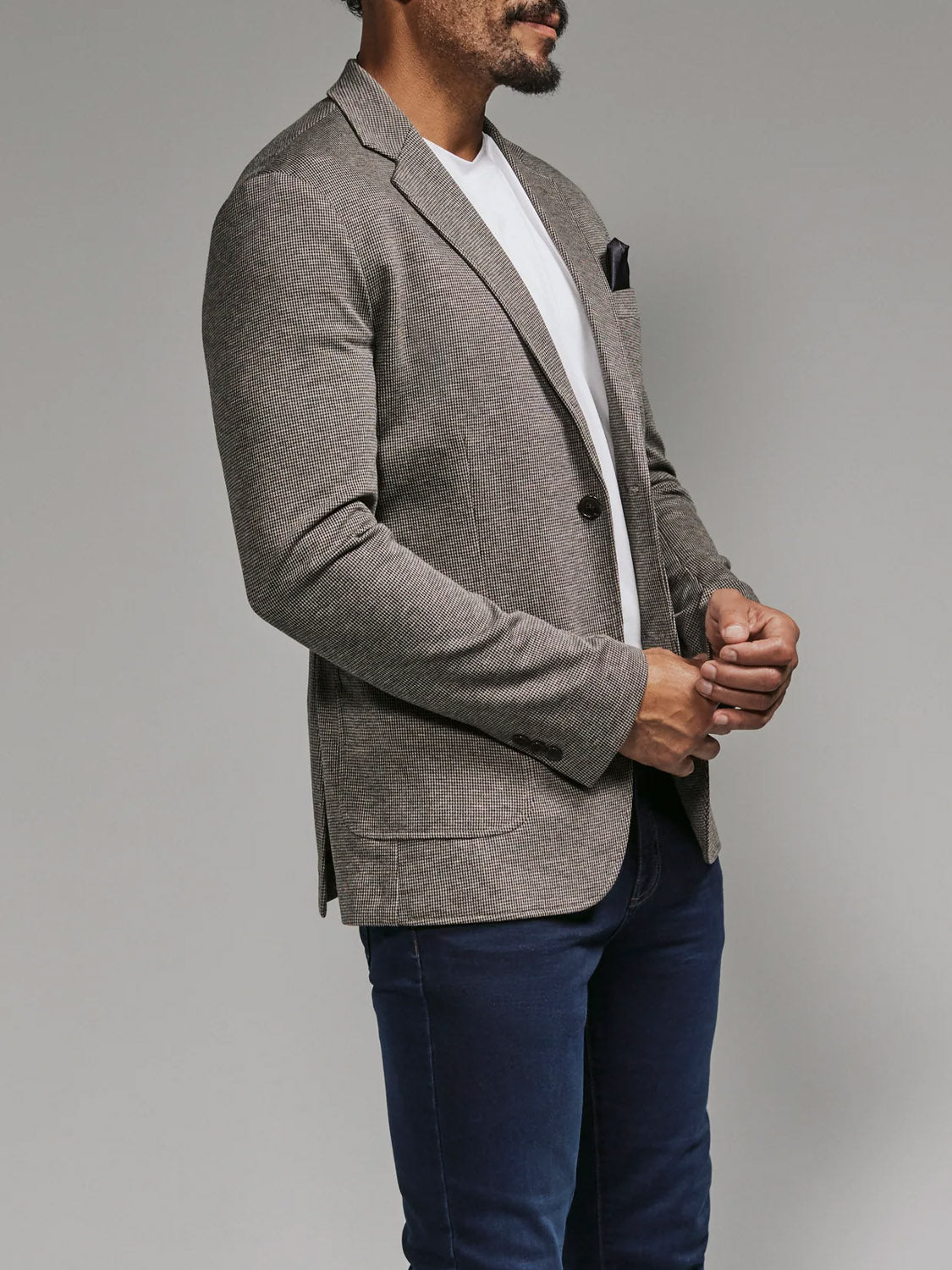 A man stands against a plain background wearing the 7 Diamonds Lafayette Blazer in gray over a white T-shirt and dark blue jeans, his hands clasped and face partially out of frame.