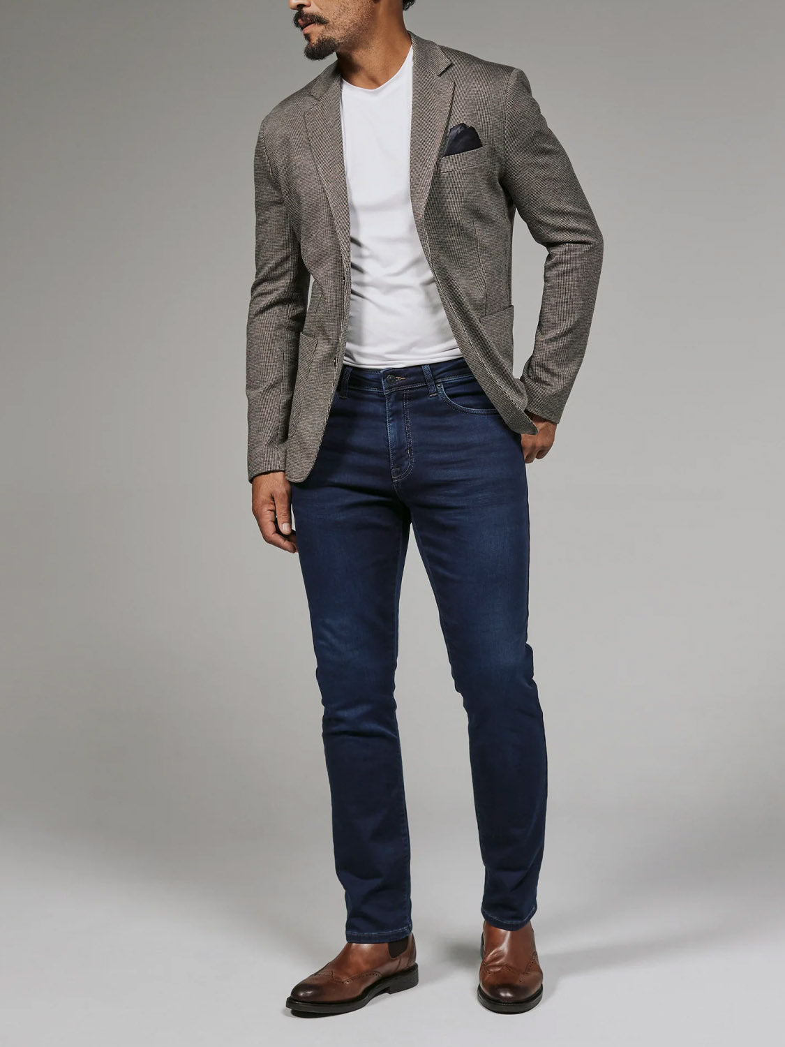 A man models the 7 Diamonds Lafayette Blazer by 7 Diamond over a white tee, paired with dark blue jeans and brown dress shoes, standing against a plain gray background with one hand in his pocket.