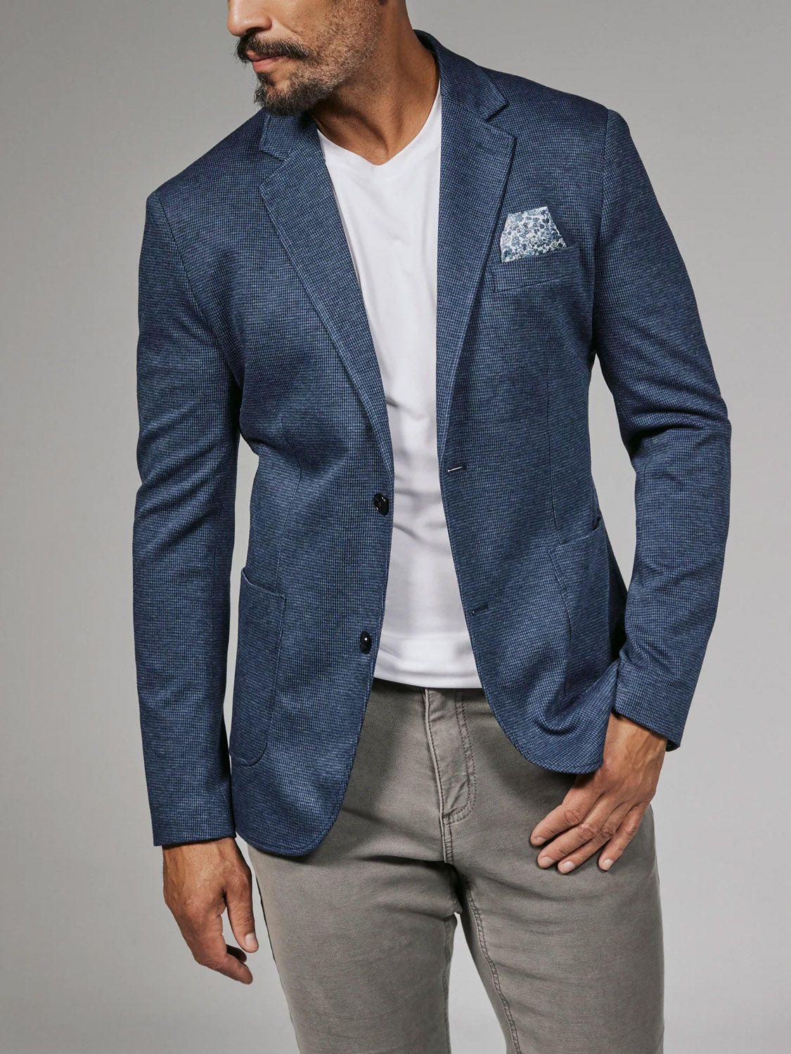 A man in the 7 Diamonds Lafayette Blazer by 7 Diamond, styled with a patterned pocket square, white t-shirt, and light gray pants, stands against a plain gray background, looking to his left.