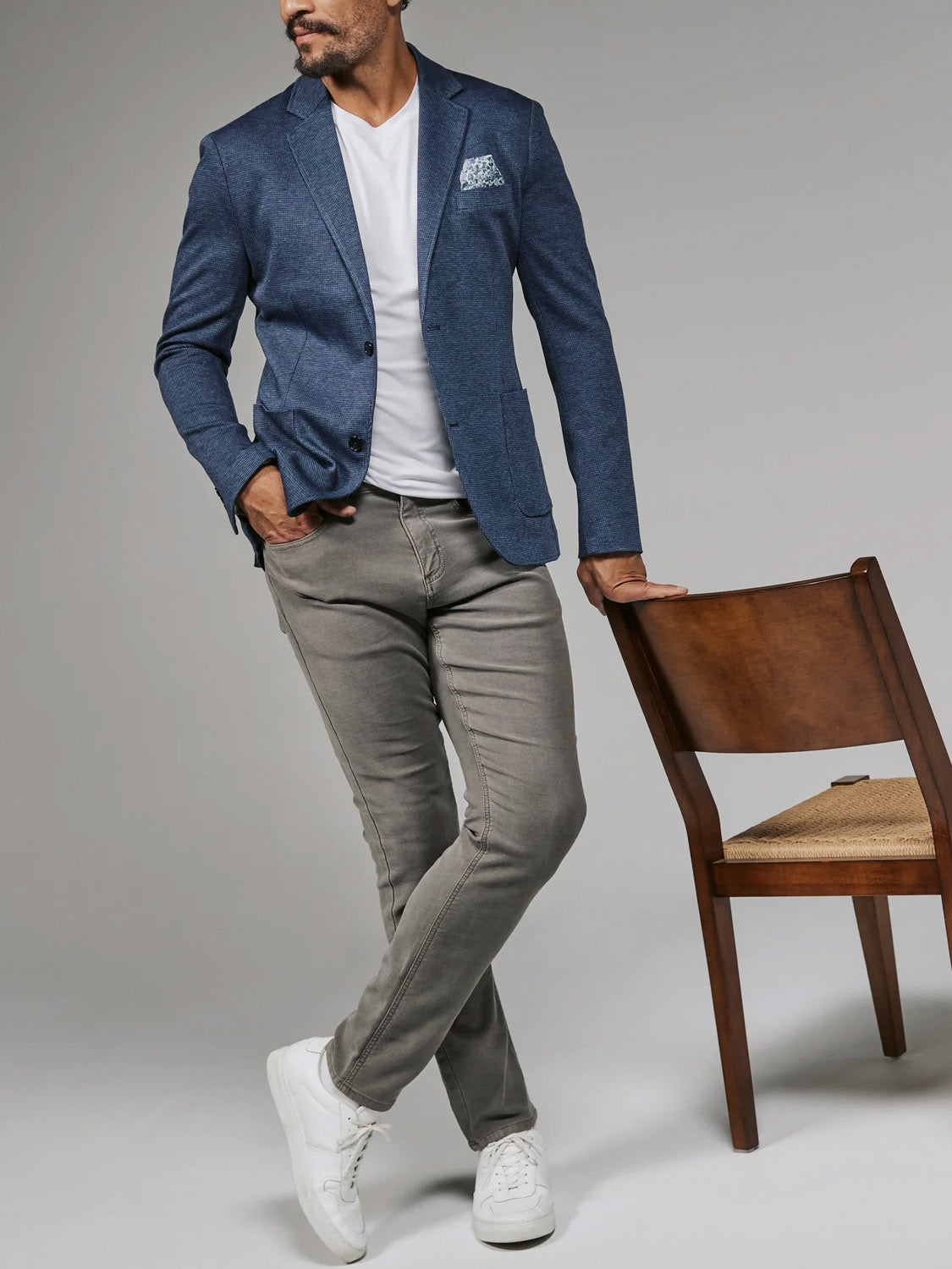 A man wearing the 7 Diamonds Lafayette Blazer in blue, a white t-shirt, gray pants, and white sneakers stands with one hand in his pocket and the other resting on the back of a wooden chair.