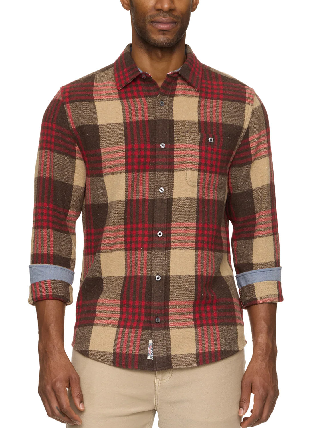 A man wears the Flag and Anthem Lansing Herringbone Shirt, featuring an athletic fit plaid in brown, tan, and red, with a chest pocket and rolled cuffs, untucked for a casual look, paired with beige pants.