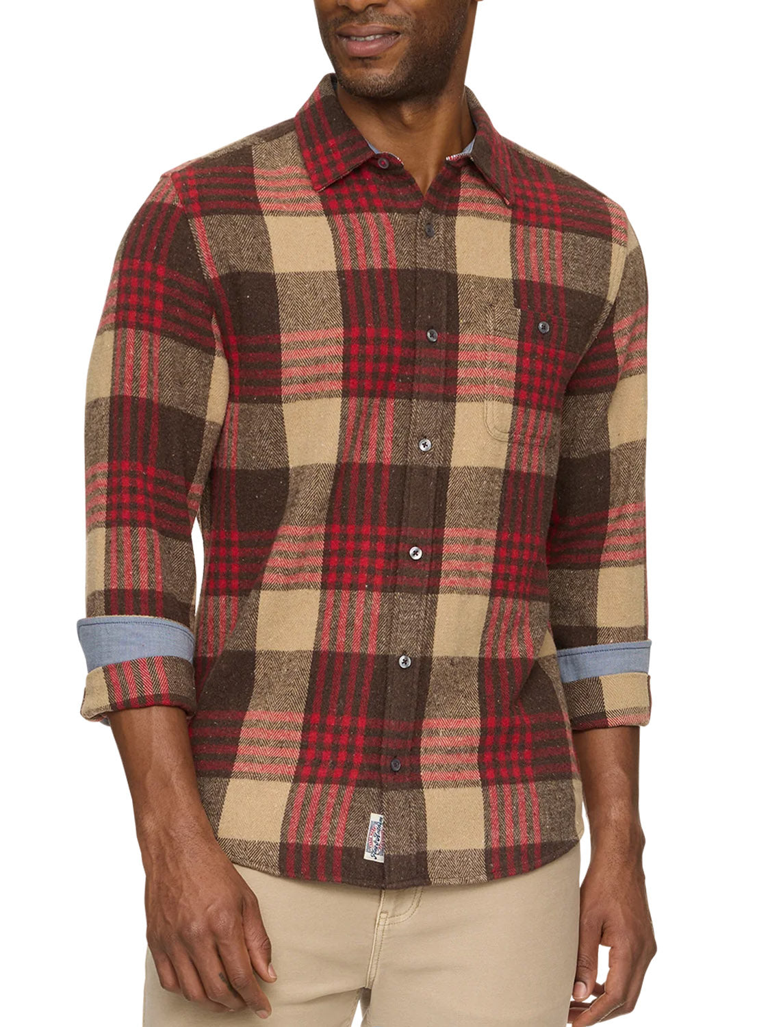A man wears the Flag and Anthem Lansing Herringbone Shirt, featuring a long-sleeve, button-up plaid design in red, brown, and beige. Its athletic fit and untucked length complement his light pants and rolled cuffs.